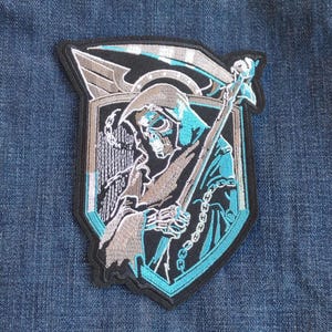 May include: Embroidered patch featuring a Grim Reaper design. The patch has a black border and a shield shape. The Reaper is depicted in shades of gray and teal, holding a scythe and chained. The background is a denim blue.