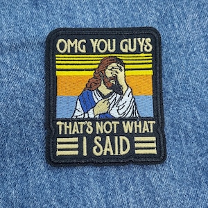 May include: A black and gold embroidered patch with a retro sunset background. The patch features a cartoon image of Jesus with his hand over his face and the text "OMG YOU GUYS THAT'S NOT WHAT I SAID".