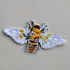 May include: Embroidered bee patch with white wings, a black and yellow striped body, and floral accents. The patch features detailed stitching and a decorative design, perfect for adding to clothing or accessories.