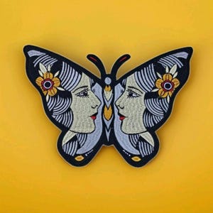 May include: Embroidered butterfly patch featuring two faces in profile, with cream-colored skin, red lips, and yellow flowers. The butterfly has a black outline and is set against a yellow background.