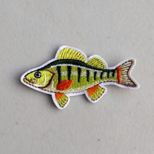 May include: Embroidered patch of a fish with yellow, green, black, and orange details. The fish has a white outline and is designed to be ironed or sewn onto fabric. The patch is a decorative item for clothing or accessories.