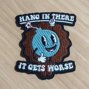May include: Embroidered patch with the text "HANG IN THERE" above and "IT GETS WORSE" below a cartoon bomb character. The bomb is blue with a smiling face and a brown explosion-like border. The patch has a black border.
