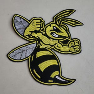 May include: An embroidered patch featuring a muscular, anthropomorphic wasp character with yellow and black coloring. The wasp has a fierce expression, clenched fists, and grey wings with black outlines, all bordered by a thick black edge.