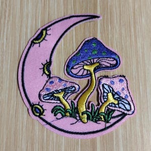 May include: Embroidered patch featuring a pink crescent moon with three colorful mushrooms. The mushrooms have blue and pink caps with yellow stems and green grass. The patch is outlined in black.