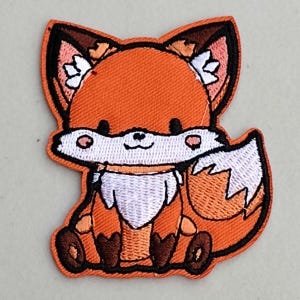 May include: A cute orange and white embroidered fox patch with a smiling face. The fox is sitting with its tail curled around its body.