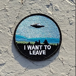 May include: A round black patch with a blue and green embroidered image of a UFO flying over a landscape. The text "I WANT TO LEAVE" is embroidered in white on the patch.