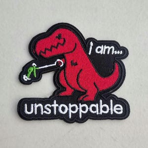 May include: Embroidered patch featuring a red Tyrannosaurus Rex holding a green figure on a white leash. The patch has the words "I am..." and "unstoppable" in white lettering on a black background.