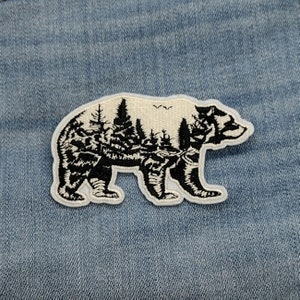 May include: A white embroidered patch of a black bear silhouette with a forest scene inside the bear's outline.