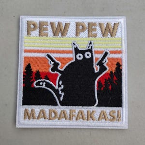 May include: Embroidered patch featuring a black cat silhouette holding two guns. The patch has the words "PEW PEW" above the cat and "MADAFAKAS!" below. The background includes a sunset design with orange, yellow, and brown stripes, and silhouettes of trees.