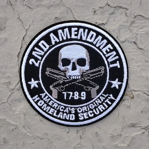 May include: Black and white embroidered patch with a skull and crossbones, two pistols, and the text "2nd Amendment", "America's Original Homeland Security", and "1789".