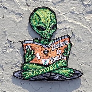 May include: Green alien reading a book with the text "Believe in Yourself". The alien is sitting in a meditative pose with a green and black background.