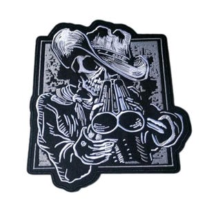 May include: A black and white embroidered patch featuring a skeleton wearing a wide-brimmed hat and a neckerchief, holding a double-barreled shotgun. The design is set against a rectangular background with a distressed, textured border pattern.