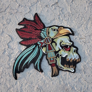May include: A colorful embroidered patch featuring a skull wearing a headdress with red, blue, and gold feathers. The skull has a fierce expression with a large, open mouth and a single red eye.