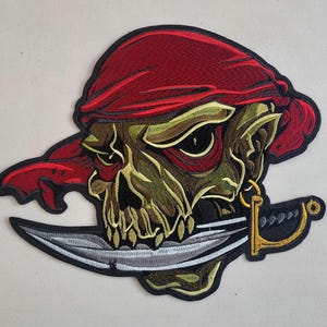 May include: Embroidered patch of a green skull wearing a red bandana and holding a silver sword. The skull has a menacing expression and is a popular design for motorcycle jackets and other apparel.