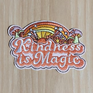 May include: Embroidered patch with the words "Kindness is Magic" in orange script. The patch features a rainbow, stars, and mushrooms in shades of orange, yellow, and pink. The patch has a white border and a black outline.