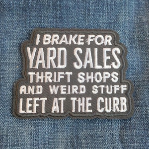 May include: A rectangular patch with a gray background and white embroidered text. The text reads "I BRAKE FOR YARD SALES THRIFT SHOPS AND WEIRD STUFF LEFT AT THE CURB". The patch has a decorative border and is set against a blue denim background.