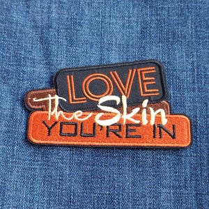 May include: Embroidered patches with the text "LOVE The Skin YOU'RE IN" in orange and white lettering. The patches are layered on a blue denim background. The top patch is navy blue, the middle patch is brown, and the bottom patch is orange.
