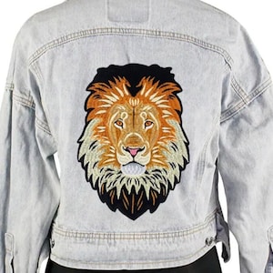 May include: Light blue denim jacket with a large embroidered lion patch on the back. The lion patch features detailed stitching in shades of orange, brown, and black, with a pink nose and a white muzzle. The jacket has a classic design.