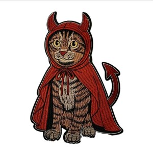 May include: An embroidered patch featuring a brown tabby cat wearing a red hooded cape with devil horns and a matching pointed tail. The cat has yellow eyes and a light-colored chest, with the entire design outlined in a thick black border.
