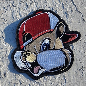 May include: Embroidered patch of a cartoon squirrel wearing a red and white baseball cap. The squirrel has a brown face, white fur, and a black outline.