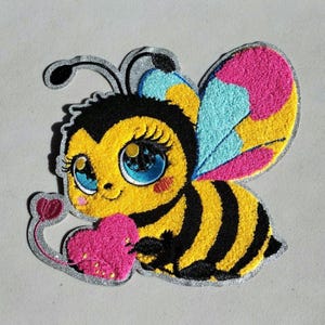 May include: A colorful embroidered bee patch with a cute design. The bee has yellow and black stripes, large blue eyes, and pink and blue wings. It holds a pink heart. The patch has a silver border.