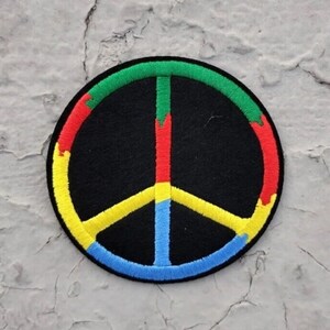 May include: A colorful peace sign patch with a black background. The peace sign is outlined in green, red, yellow, and blue.