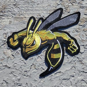 May include: A yellow and black embroidered patch of a cartoon wasp with its wings spread out. The wasp is in a flying position with its stinger pointed forward. The patch is made of felt and has a black border.