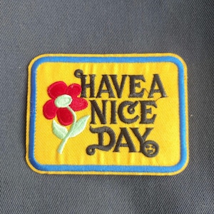 May include: Embroidered patch with a yellow background, blue border, and the words "HAVE A NICE DAY" in black script. A red flower with green leaves is on the left side. The patch is rectangular.
