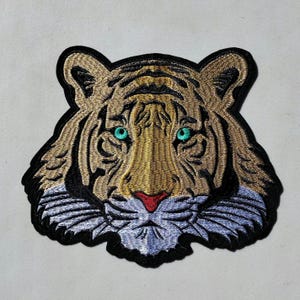 May include: Embroidered patch of a tiger's face with a black outline, brown and gold fur, and bright turquoise eyes.