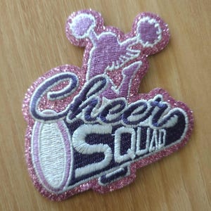 May include: A glittery, embroidered patch with the words "Cheer Squad" in purple and white. The patch features a cheerleader with pom-poms and a pink glitter border. The patch is on a wooden surface.