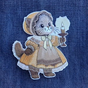 May include: Embroidered patch featuring a vintage-style cat wearing a bonnet and dress, holding a candle. The patch has a detailed design with brown, tan, and yellow colors, and a white border. The patch is likely for clothing or accessories.