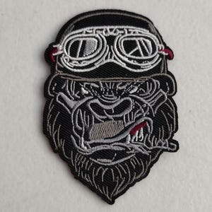 May include: Black and white embroidered patch of a gorilla wearing goggles. The gorilla has a fierce expression and is smoking a cigarette.