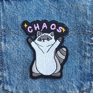 May include: An embroidered patch featuring a white and gray raccoon with its arms raised, positioned below a black speech bubble containing the pink text "CHAOS" and two yellow stars, all attached to a blue denim fabric background.
