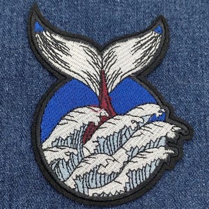 May include: Embroidered patch of a white whale tail with black outline, emerging from blue water with white waves. The patch is on a denim background.