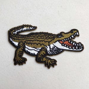 May include: Embroidered patch featuring a detailed crocodile design. The patch showcases a green and brown crocodile with a white underbelly and open mouth revealing red accents. The edges are outlined in black.