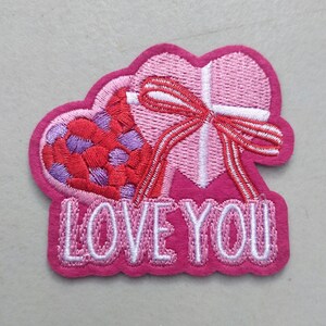 May include: A pink embroidered patch featuring two hearts and a bow. One heart is filled with red and purple dots, the other is a pink heart with a white bow. The words "LOVE YOU" are embroidered in white.