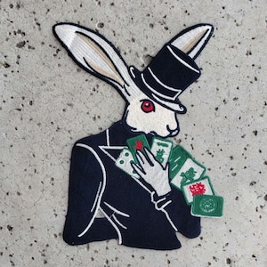 May include: A white rabbit wearing a black top hat and a blue suit holds a hand of playing cards. The cards are green with red accents. The rabbit has a red eye.