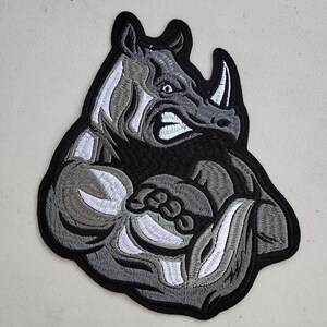 May include: A black and white embroidered patch of a muscular rhinoceros with a fierce expression. The rhinoceros is standing on its hind legs with its front legs crossed. The patch is perfect for adding a touch of personality to jackets, bags, or other items.