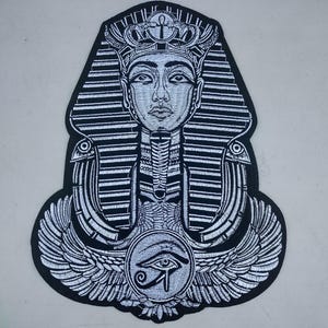 May include: A black and white embroidered patch featuring the stylized image of an Egyptian pharaoh. The pharaoh is depicted with a detailed headdress, a serene facial expression, and a prominent eye symbol at the bottom.
