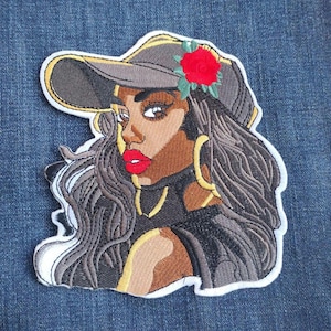 May include: Embroidered patch featuring a stylized portrait of a woman wearing a gray baseball cap adorned with a red rose. The woman has long, wavy hair, gold hoop earrings, and red lipstick. The patch has a white border and is set against a denim background.