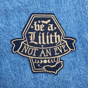 May include: A navy blue embroidered patch with the text "be a Lilith NOT AN EVE" in an ornate font. The patch has a coffin-like shape with bats and moon phases. The patch is on a blue denim background.