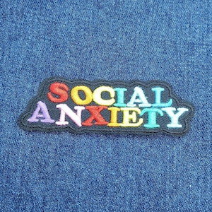 Puede incluir: Parche bordado con las palabras "SOCIAL ANXIETY" en letras coloridas. El parche tiene un borde negro y está sobre un fondo de mezclilla azul. Las letras son rojas, amarillas, turquesas, moradas y naranjas.