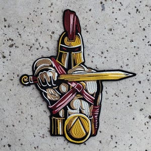 May include: Embroidered patch of a golden Spartan warrior with a red plume on his helmet, holding a sword and shield with a golden triangle design.