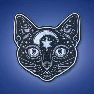 May include: Black cat face patch with white embroidered details. Features a crescent moon, star, and celestial accents. The cat's eyes and whiskers are also outlined in white. The patch is set against a dark blue background.