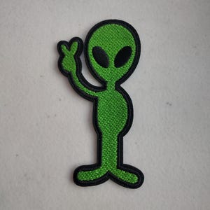 May include: Embroidered green alien patch with black outline. The alien is depicted with large black oval eyes and is giving a peace sign with its hand. The patch is made of a textured fabric and is likely designed to be ironed or sewn onto clothing or accessories.