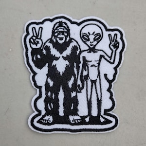 May include: Embroidered patch featuring a black and white design of a Bigfoot and an alien, both making peace signs. The patch has a white border and is likely for clothing or accessories.