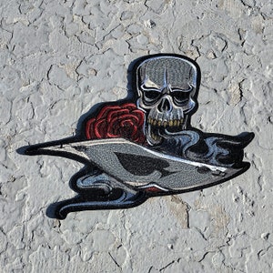 May include: A silver skull with a red rose and smoke coming out of its mouth, embroidered on a black patch.