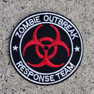 May include: A black and white embroidered patch with a red biohazard symbol in the center. The text around the symbol reads "Zombie Outbreak Response Team".