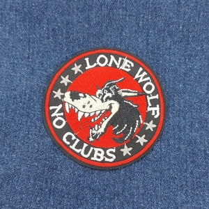 May include: A black and white embroidered patch with a red background. The patch features a cartoon wolf with a toothy grin and the text "Lone Wolf No Clubs" in a circle around the wolf.