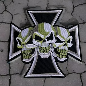 May include: A black iron cross with three white and green skulls embroidered on it. The skulls have a striped pattern and are facing forward.